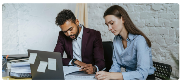 Two people sit at a desk covered in papers engaged in an editing process.